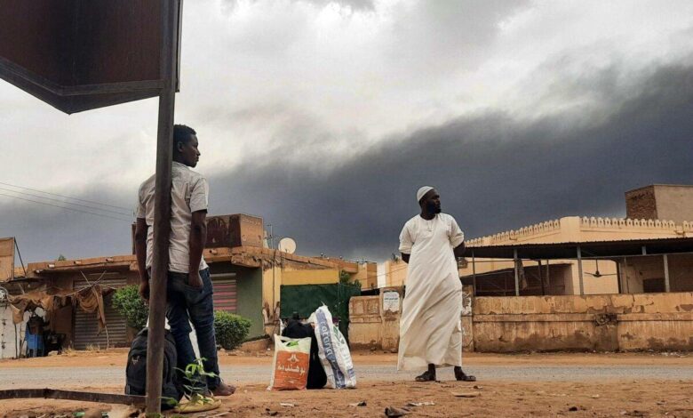 Smoke rises above buildings as people wait on the side of a road with some belongings, in Khartoum