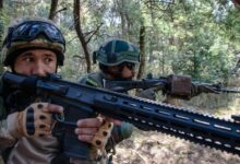 Two soldiers are seen patrolling in what appears to be a forest in daylight with their rifles at the ready. Both of them are pointing their weapons to the right. The one on the foreground, positioned on the left, is holding a Sig Sauer rifle. The other soldier is positioned behind his left, on the center of the image. They are both wearing camouflage.