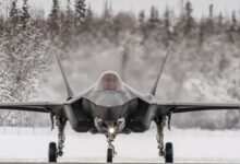 A black F-35 fighter jet plane is seen grounded, facing the viewer. The area it is in is snowy, with the ground completely covered in white snow. The background is a forest of snow-covered trees. The background on the back of the plane is blurred, presumably due to the heat distortion emitted by the plane.