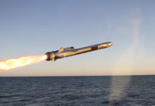 Kongsberg's Naval Strike Missile (NSM) is seen front and center, launching from the left. A jet of red-orange flame streaks from its back. The background is a deep blue ocean, with a cloudless sky farther back.