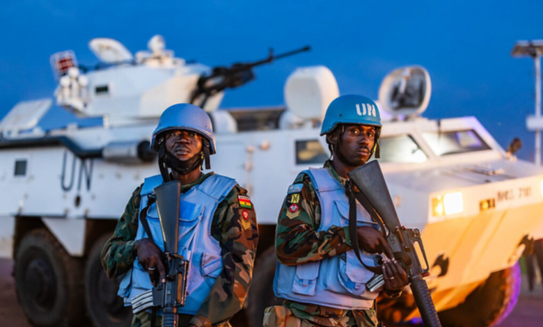Two uniformed soldiers stand front and center of the image, staring straight. They both hold automatic rifles, pointed down. They are wearing a blue helmets and vests. The soldier on the right has 'UN' written in white in front of his helmet. Underneath their vests, they have a camouflage uniform on. A white tank with 'UN' written on its body in black is seen stationed in the background. Its orange headlights are on. The background is a dark blue sky.