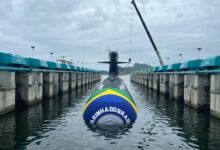 The Tonelero (S42), Brazil's third Riachuelo-class submarine, is seen half-submerged in a pier. Its front is wrapped in clothing with Brazil's colors, as well as the Brazilian Navy's logo in the center. The background is a dull sky covered in dark clouds.