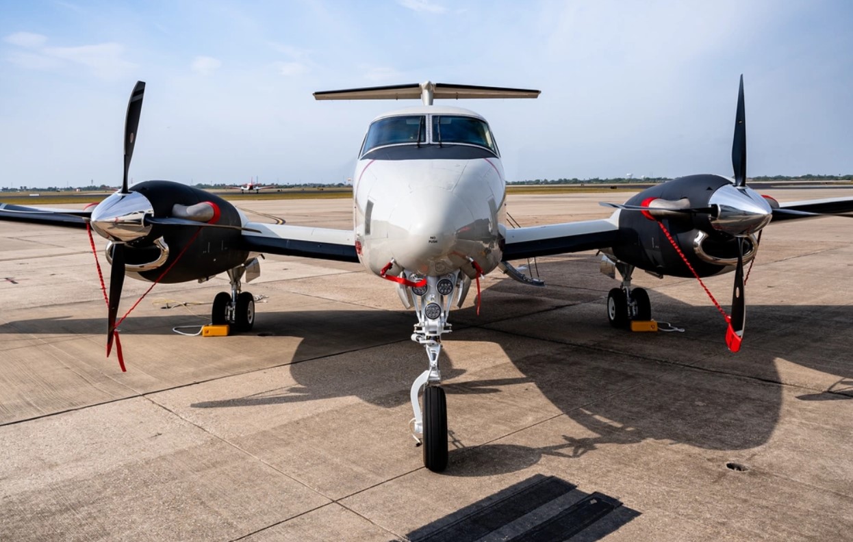 Textron's T-54A multi-engine training system aircraft is seen parked in the middle of what appears to be an aircraft runway. The plane has two propellers, one on each of its wings. The aircraft is painted white with navy blue accents on its wings and the front of the cockpit. The background is a clear blue sky with a few streaks of light, white clouds.