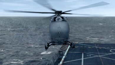 A UH-72 Unmanned Logistics Connector is seen hovering above a helicopter deck in the middle of the sea. The helicopter is painted dark silver gray.