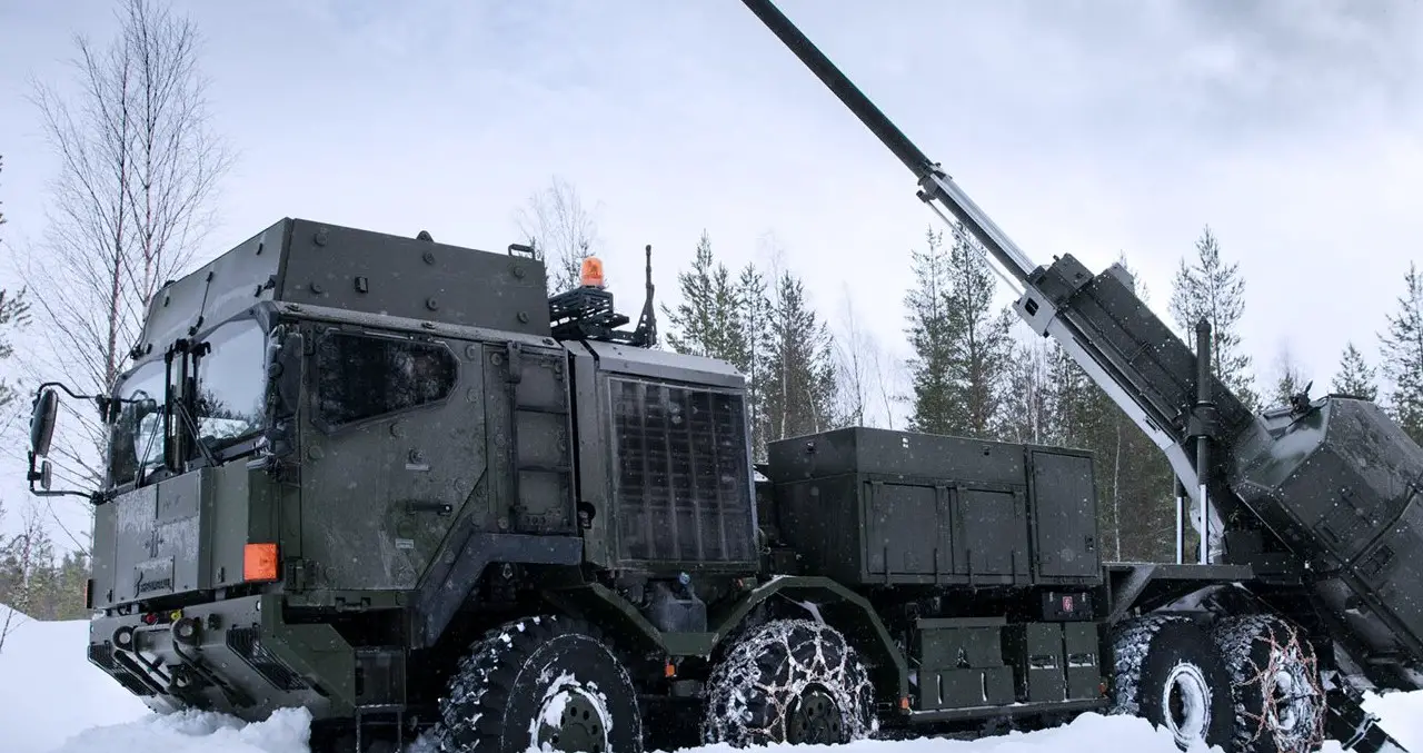 A Rheinmetall HX truck is seen parked on a snowy field. The grayish-green 8x8 truck has chains fitted on some of its tires to help with traction. On its back, a large turret is attached and pointing northwest. The ground is covered completely by snow, covering almost the entire bottom half of the truck's wheels. The background is a sparse group of thin trees with little to no leaves, and a gloomy, white sky.