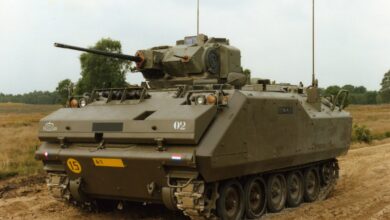 A YPR-765 armored infantry fighting vehicle is seen stationed on a dirt road. The vehicle has the typical appearance of a military tank, with a boxy built, a turret, and tracked wheels. The vehicle is painted grayish green. The background is a horizon of green trees and a clear white sky.