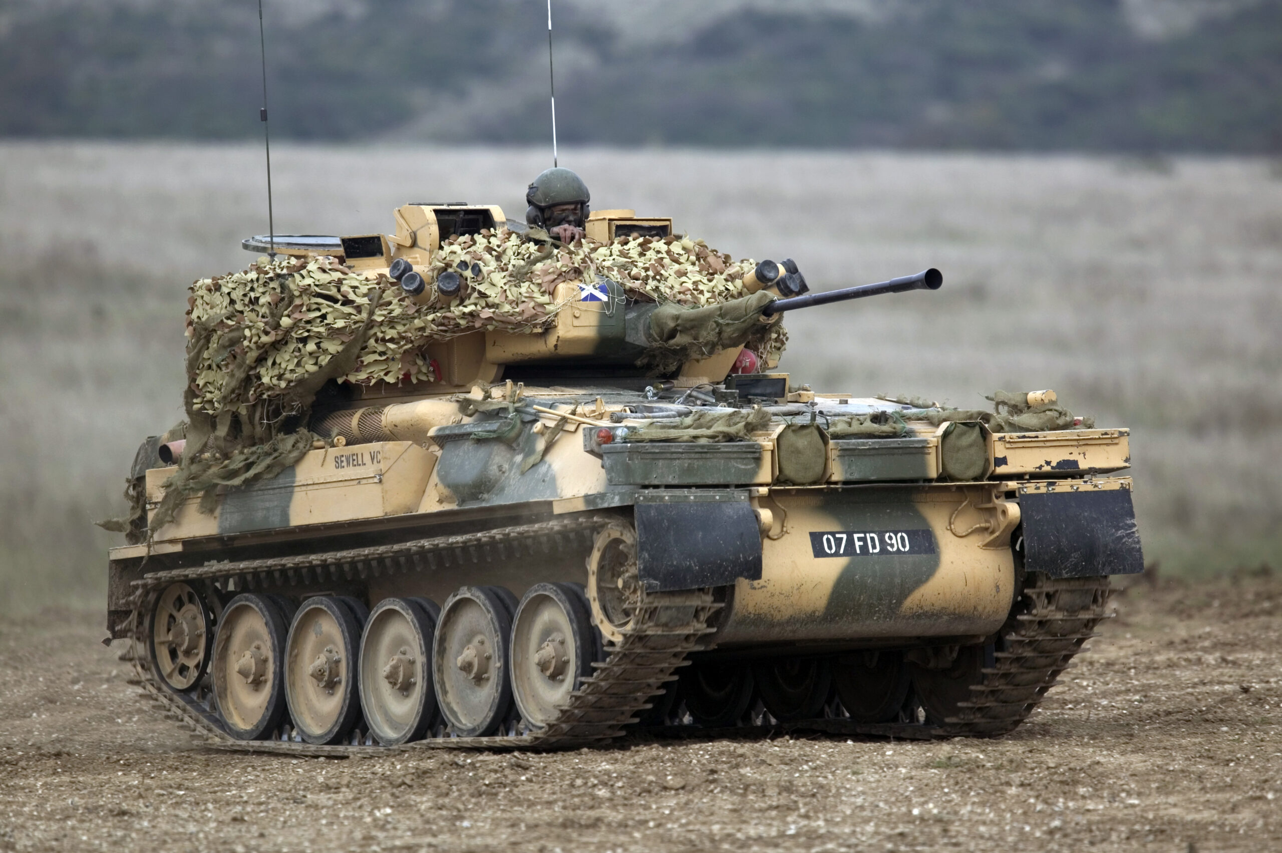 An FV107 Scimitar armored tracked reconnaissance vehicle fleet is seen in the middle of a plains. The vehicle is painted camouflage brown and green. A soldier wearing protective goggles and a green helmet is seen peeking out from the vehicle's hatch. The background is out of focus but appears to be a forested area.