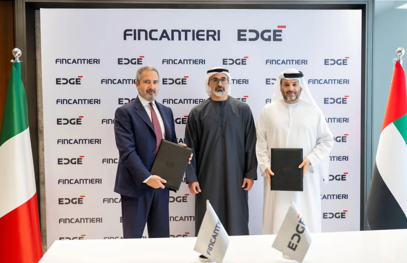 Fincantieri CEO and Managing Director Pierroberto Folgiero, Abu Dhabi crown prince Khaled bin Mohamed Al Nahyan, and EDGE Group chairman Faisal Al Bannai stand to take a photo op during a signing ceremony. Folgiero and Al Bannai hold leather dosiers, which may be holding the signed documents. Italy's flag is seen displayed on the left, and UAE's is on the right. On a white table in front of them, two flags printed with Fincantieri and EDGE's logos are also propped up. Behind them is a white screen with the companies' logos printed in a repeating pattern.