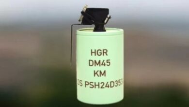 A Rheinmetall DM 45 hand grenade is displayed. The grenade has a cylindrical shape and is painted green, with "HGR DM45 KM" and a long serial number inscribed on it.