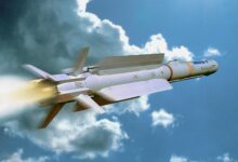 An IRIS-T missile is seen flying in a cloudy blue sky. It juts out smoke and red and white flames as it propels upward. The missile is sleek, painted gray, and has four fins attached to its bottom.