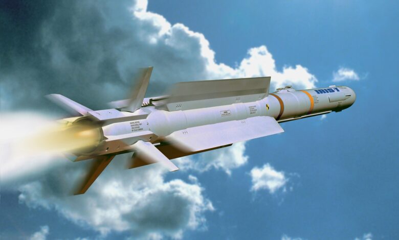 An IRIS-T missile is seen flying in a cloudy blue sky. It juts out smoke and red and white flames as it propels upward. The missile is sleek, painted gray, and has four fins attached to its bottom.