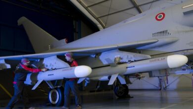 Two gray MBDA Marte ER missiles are seen attached to a similarly-painted jet plane parked inside what appears to be a hangar. Two workers inspect one of the missiles set underneath the plane's right wing.