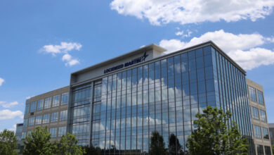 An image of the new Lockheed Martin facility in Huntsville, Alabama. The building has glass panels for its front, which reflects the blue sky dotted with white clouds. Some trees can be seen peeking out from the foreground.