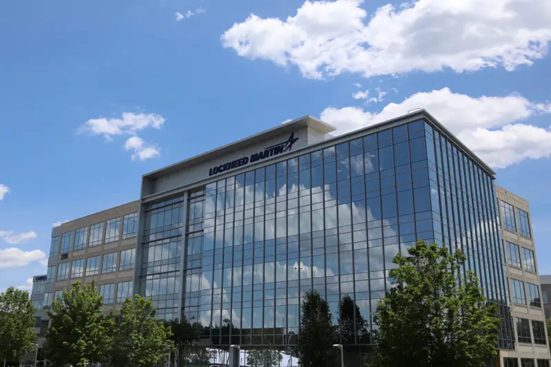 An image of the new Lockheed Martin facility in Huntsville, Alabama. The building has glass panels for its front, which reflects the blue sky dotted with white clouds. Some trees can be seen peeking out from the foreground.