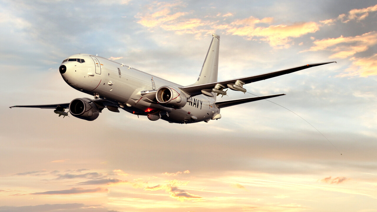 A US Navy P-8A Poseidon aircraft is seen flying in a dusk sky illuminated by the sun's orange light.