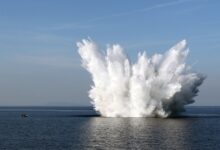 A naval mine sets off in the middle of an ocean. The water's white mist is seen blasting off on all directions. On the far left side of the image, a small boat is seen.