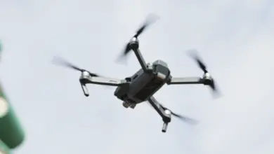 A black drone is seen hovering above. It has four propellers, each of them attached to the end of its four "limbs". The background is a gloomy sky covered with dark white clouds.