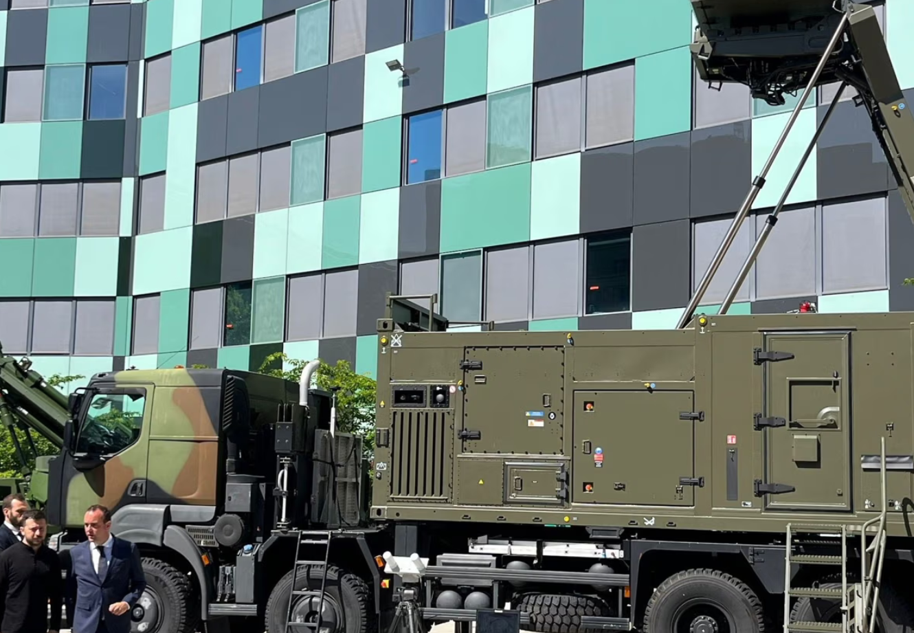 A Thales ControlMaster 200 is seen displayed in front of a building with window panes in gray, light green, and dark green colors. The system's truck is painted in deep green and brown camouflage colors. Ukrainian president Volodymyr Zelenskyy is seen walking on the bottom left side of the image with two other people wearing suits.