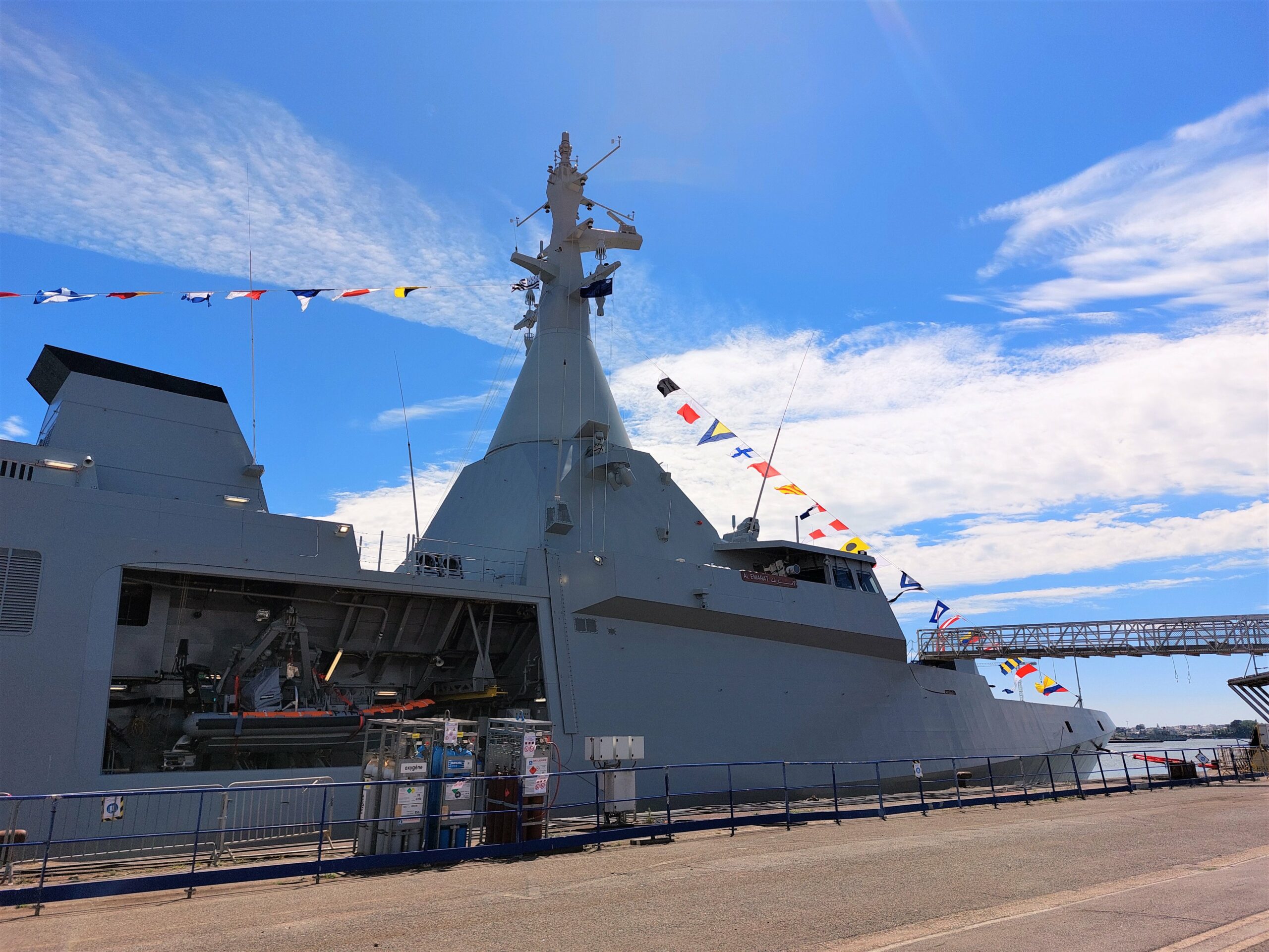 Al Emarat, the UAE Navy's second Gowind-class corvette, is seen docked at a pier. The ship is decorated with colors and flags attached to a banner hoisted up the ship's mast.
